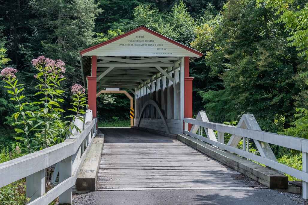 A historic red and white covered bridge spans a quiet road in Bedford County, Pennsylvania, surrounded by lush greenery and wildflowers.